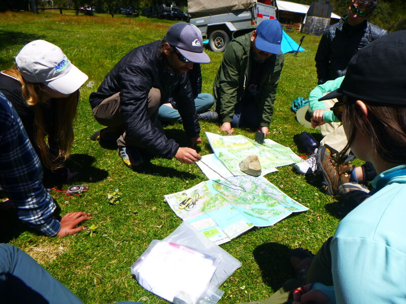 A group of people are gathered on a grassy field, seemingly engaged in a map-reading activity. They are huddled around several maps spread out on the ground, with some individuals pointing and discussing. The setting appears to be outdoors, possibly a park or recreational area, with a vehicle and trailer visible in the background. The atmosphere suggests a collaborative and educational environment.